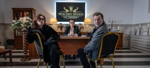 Trzy osoby siedzą w eleganckim biurze domu pogrzebowego. W tle logo "Wieczny Spokój". Scena z serialu "Minuta ciszy sezon 2".