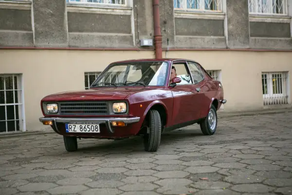Czerwony fiat 128, klasyk z lat 70., zaparkowany na brukowanym dziedzińcu przed budynkiem.
