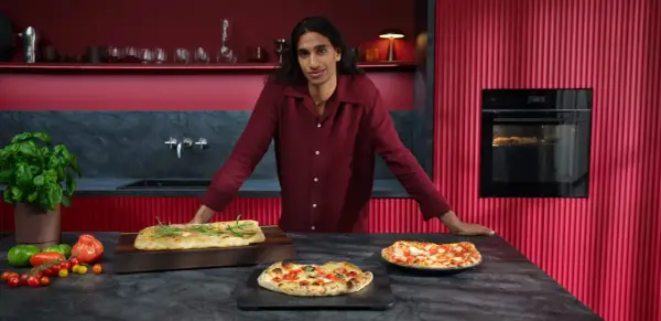 Mann in bordeauxrotem Hemd präsentiert frisch gebackene Pizzen. Im Hintergrund ein Ofen mit Pizzafunktion, der eine weitere Pizza bäckt.