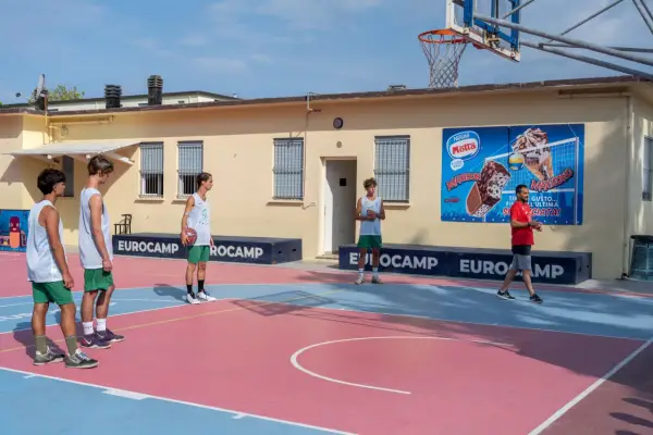 Ragazzi in tenuta sportiva giocano a basket in un camp estivo a Cesenatico, con un cartellone pubblicitario di gelati Motta sullo sfondo.