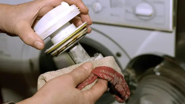 Hände reinigen das Flusensieb der Waschmaschine. Ein Tuch wischt Schmutz von dem ausgebauten Teil.