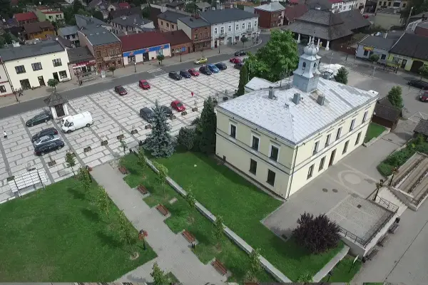 Sławków rynek z lotu ptaka: żółty budynek ratusza, brukowany plac z samochodami i zielony park z ławkami.