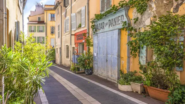 Bunte Häuser und Pflanzen säumen eine Gasse in der Marseille Altstadt. Ein "Bazar du Panier" Schild und blaue Holztüren fallen auf.