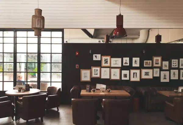 Una lámpara de yute cuelga sobre mesas y sillones de cuero en un café con paredes oscuras y una gran ventana.