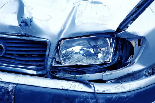 Il cofano e il faro di un'auto sono gravemente danneggiati, con lamiera accartocciata e vetro rotto. Sembra il risultato di **atti vandalici**.