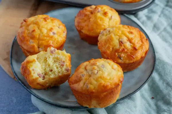 Fünf herzhafte Muffins mit Speck und Käse auf einem grauen Teller. Einer ist angeschnitten und zeigt die saftige Krume.