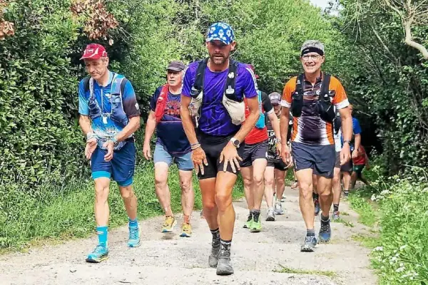 Coureurs en tenue de trail, certains avec des sacs d'hydratation, sur un sentier boisé. Le coureur au centre, portant une casquette bleue, semble en pleine course, peut-être en quête d'un record gr34.