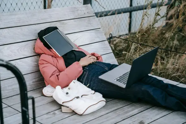 Osoba w różowej kurtce leży na ławce, z zeszytem na twarzy. Obok leży laptop i plecak. Czas na odpoczynek i dodanie nowych pomysłów.