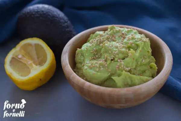 Hummus di avocado cremoso in ciotola di legno, guarnito con semi di sesamo. Accanto, un avocado intero e mezzo limone.