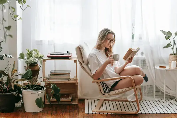 Mujer leyendo en un sillón, taza en mano, rodeada de plantas y libros. Un rincón de lectura con decoración acogedora y un tocadiscos.