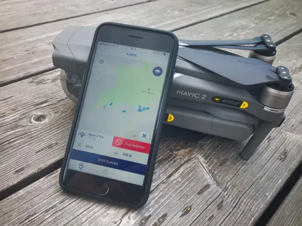 Eine Drohne liegt neben einem Smartphone, auf dem eine Drohnen-App mit Karte und Flugverboten angezeigt wird.