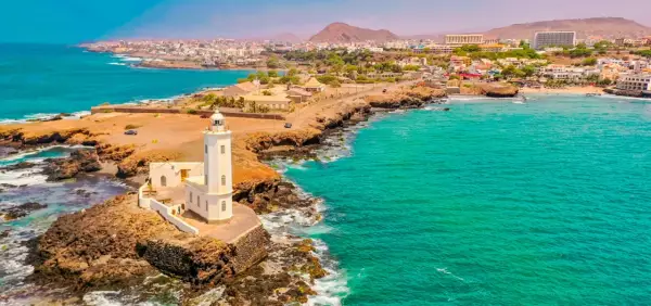 Faro blanco en una costa rocosa con aguas turquesas. Un pueblo costero se extiende detrás. ¡Ideas de qué hacer en Cabo Verde!