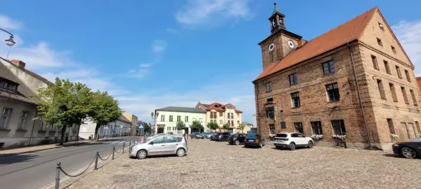 Rynek w Połajewie z zabytkowym ratuszem i kolorowymi kamienicami. Na brukowanym placu zaparkowane samochody.