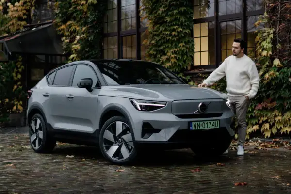 Szary Volvo C40 z czarnym dachem stoi na mokrej, brukowanej nawierzchni. Mężczyzna w białym swetrze opiera się o maskę.