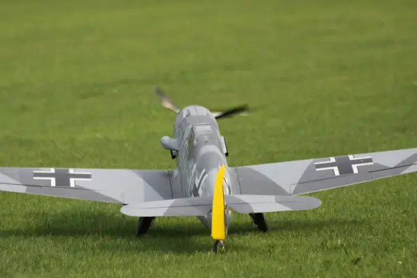 Modellflugzeug einer Me109 mit Tarnmuster und gelbem Seitenleitwerk auf einer grünen Wiese.