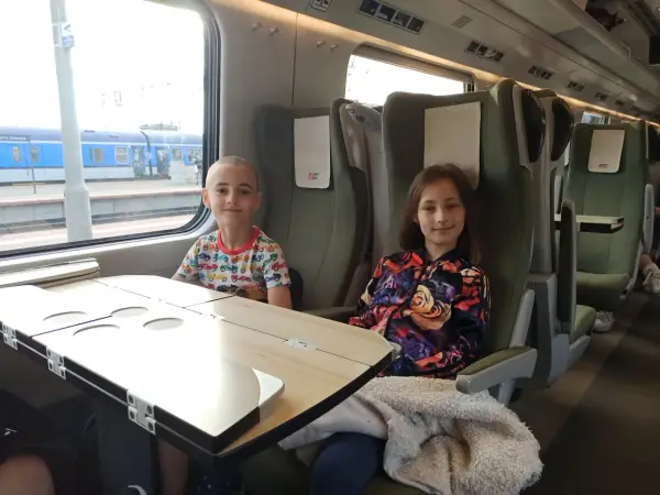Dzieci siedzą w przedziale dla osoby z dzieckiem do lat 6 Intercity. Chłopiec w kolorowym T-shircie i dziewczynka w kurtce z kwiatowym wzorem.