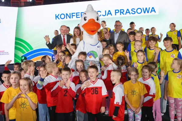 Uśmiechnięte dzieci i duża maskotka z logo lekkoatletycznych mistrzostw świata. Wszyscy pozują do zdjęcia, w tle napis "Konferencja prasowa".