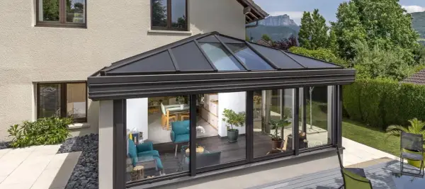 Une **pergola bioclimatique** moderne avec toit vitré, prolongeant une maison vers un jardin verdoyant. L'intérieur lumineux révèle un salon cosy et une salle à manger.