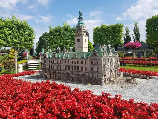 Miniatura ratusza z wieżą zegarową, otoczona czerwonymi kwiatami, w parku miniatur w Kotlinie Kłodzkiej.