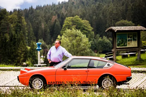 Ein Mann lehnt an seinem leuchtend orangen Simca Matra Bagheera. Im Hintergrund ein dichter Nadelwald.