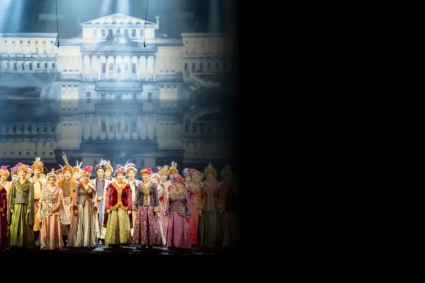 Chór w barwnych strojach historycznych na tle pałacowej scenografii. Sprawdź opera warszawa repertuar!