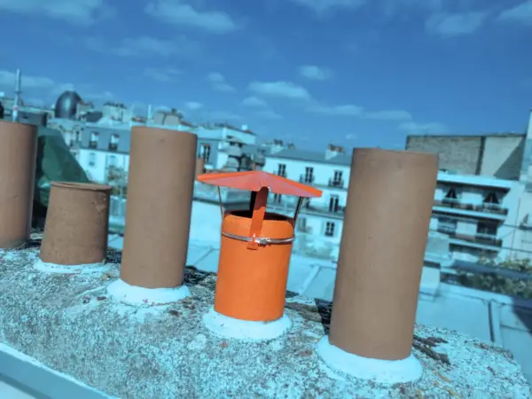 Sur un toit parisien, un chapeau chinois orange protège une cheminée, entouré d'autres conduits sous un ciel bleu.