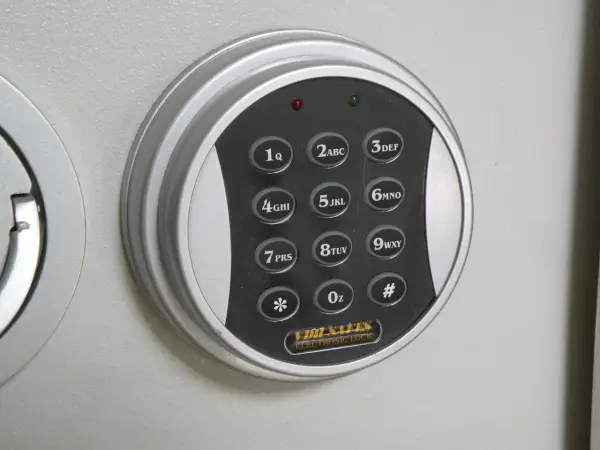 Elektroniczna klawiatura szyfrowa sejfu z przyciskami cyfrowymi i symbolami. W tle widoczne są metalowe tuleje mocujące.