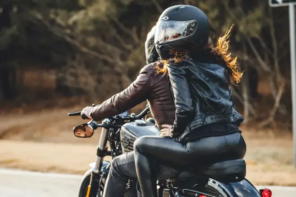 Coppia in moto, lei indossa giacca di pelle e pantaloni neri, lui giacca marrone. Perfetto esempio di come vestirsi per andare in moto passeggero.