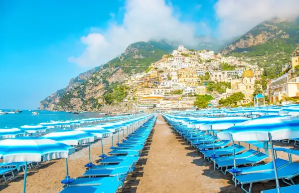 Rzędy leżaków i parasoli na plaży w Positano, Włochy. W tle widać malownicze miasteczko na zboczu góry i błękitne morze.