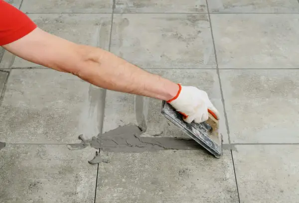 Mano con guante aplica lechada gris a las juntas entre azulejos de cemento con una llana.