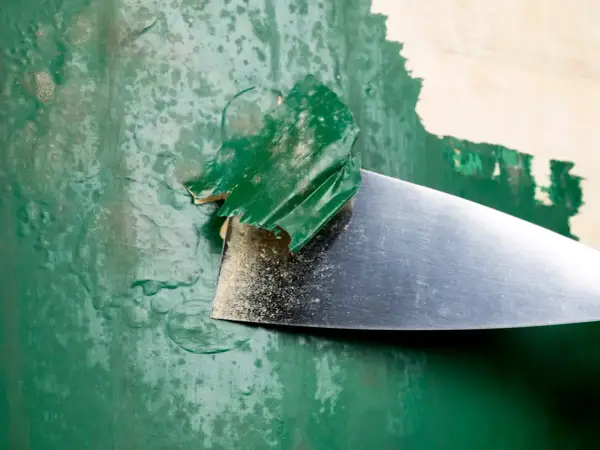 Metalowa szpachelka zdziera starą, zieloną farbę, odsłaniając podkład.