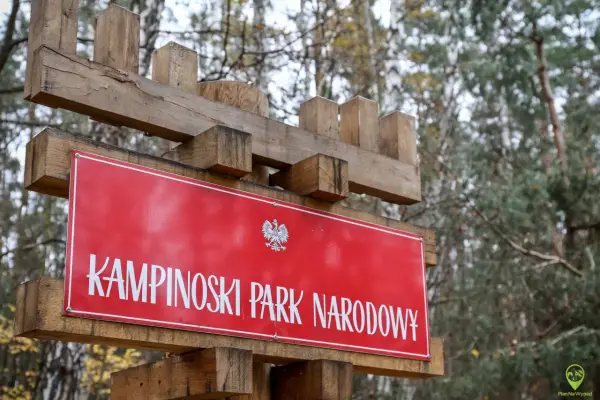 Drewniany drogowskaz z napisem "KAMPINOSKI PARK NARODOWY" i godłem Polski. W tle las.