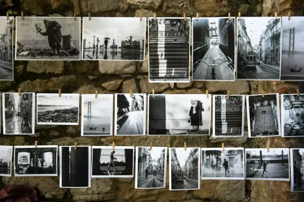 Czarno-białe fotografie wiszą na sznurku na kamiennej ścianie. Widać na nich uliczne sceny z Lizbony, w tym tramwaje, most i ludzi.
