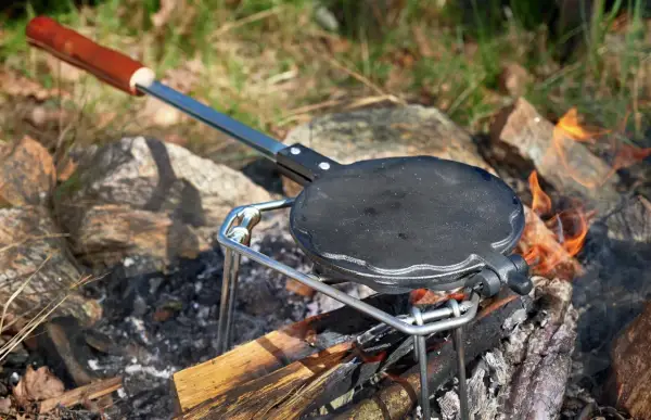 En gjutjärnspanna med trähandtag står på en ställning över en öppen eld. Perfekt för att steka plättar eller våfflor ute i naturen.