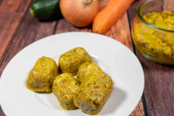 Cinque cubetti dorati di dado vegetale fatto in casa, preparati con carote, zucchine e cipolle, su un piatto bianco.