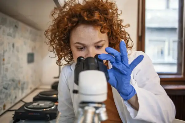 Badania do hematologa: młoda kobieta w niebieskich rękawiczkach bada próbkę pod mikroskopem.