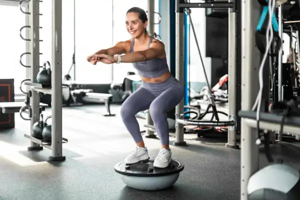 Frau macht Kniebeugen auf einem Bosu-Ball, trägt Sportkleidung und weiße Sneaker. Sie trainiert ohne schuhe auf dem instabilen Untergrund.