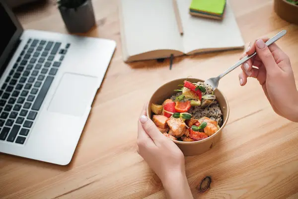 Ręce trzymające miskę z łososiem i warzywami, obok laptop i notatnik. Idealny posiłek na dieta okienkowa.