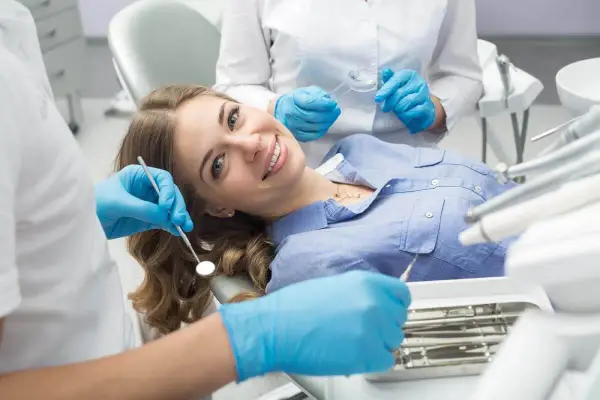 Uśmiechnięta pacjentka na fotelu dentystycznym, mimo że trochę się dentysty boi. Asystentka przygotowuje narzędzia.