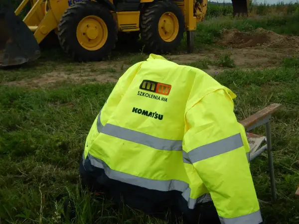Kamizelka z logo Komatsu i napisem "Szkolenia MM" leży na krześle obok koparki. Idealna na testy na koparki jednonaczyniowe.