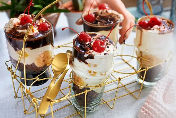 Schokoladige Dessert-Rezepte in Gläsern mit Bananen, Sahne und Kirschen. Goldene Löffel liegen bereit.