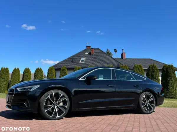 Elegancki czarny Audi A7 C8 lśni w słońcu na tle domu i zielonych drzew.