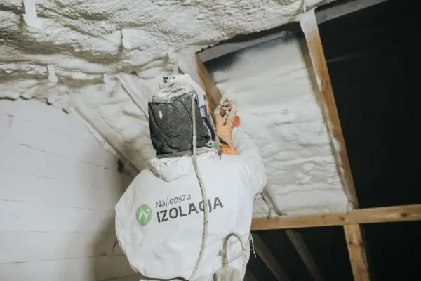 Pracownik w kombinezonie natryskuje twardą piankę poliuretanową na poddasze, tworząc skuteczną izolację.
