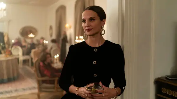 Alicia Vikander i en elegant svart klänning med stora örhängen, håller ett glas. Scenen utspelar sig på en tillställning, kanske från en av hennes filmer.