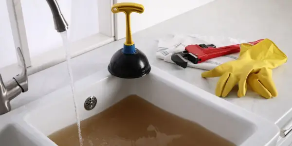 Un évier bouché après siphon, avec de l'eau sale stagnante. Un déboucheur, une clé à molette rouge et des gants jaunes sont prêts à l'emploi.
