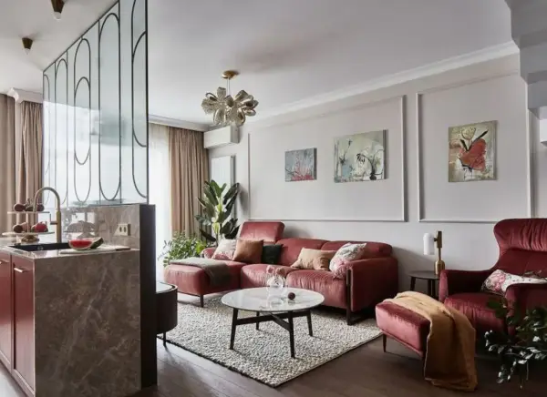 Elegancki salon z bordową kanapą i fotelem. Modne ściany w salonie zdobią obrazy, a kuchnia oddzielona jest ażurową ścianką.