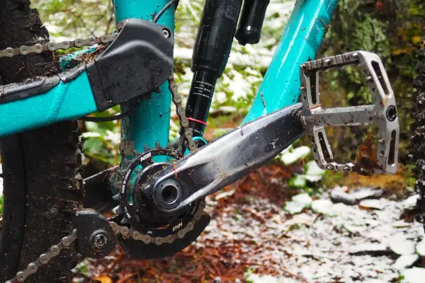 Close-up of a muddy mountain bike drivetrain with a teal frame, showing the crankset, chain, and pedal.
