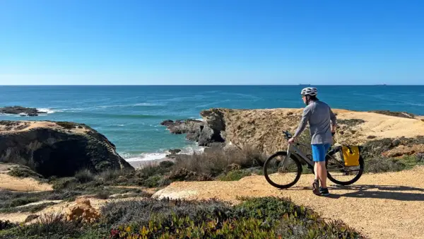 A cyclist pauses on a cliffside path during a Porto to Lisbon bike tour, admiring the vast blue ocean.
