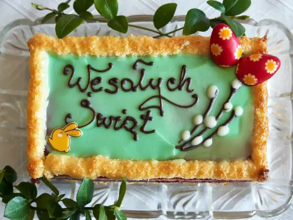 Zielone ciasto wielkanocne z napisem "Wesołych Świąt", ozdobione zajączkiem, baziami i pisankami.