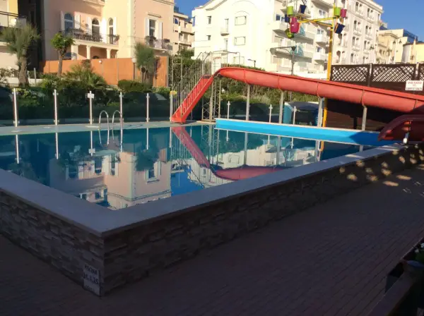 Piscina con scivolo rosso e giochi d'acqua ai Bagni Carmen Cattolica. Riflessi degli edifici nell'acqua azzurra.